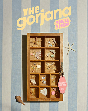 Display of shell-themed jewelry in a wooden box with 'The Gorjana Shell Shop' branding on a striped background.